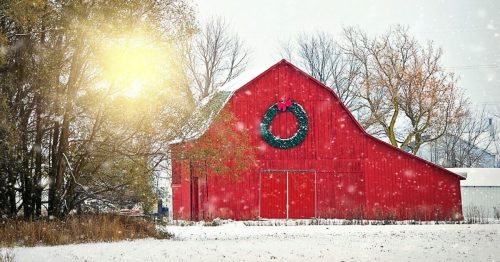A red barn decorated with a wreath, surrounded by snow and trees, with soft sunlight filtering through the falling snow.