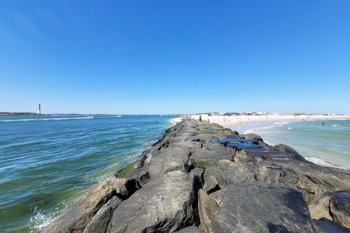 A rocky jetty extends into a calm blue sea, with a sandy beach and clear sky in the background.