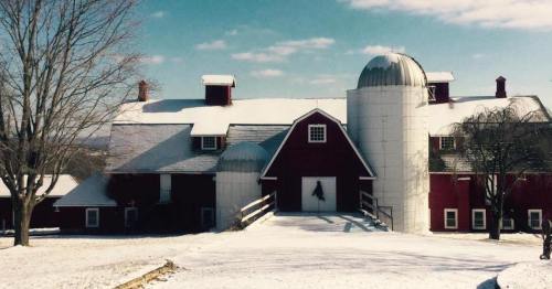 A red barn and silos covered in snow, set against a clear blue sky and winter landscape.