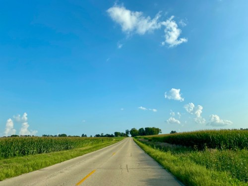 A straight road stretches into the distance, flanked by green fields and a clear blue sky with scattered clouds.