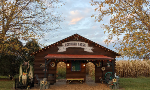 A rustic wooden barn labeled "Reindeer Ranch," surrounded by autumn trees and cornfields, with festive lights.