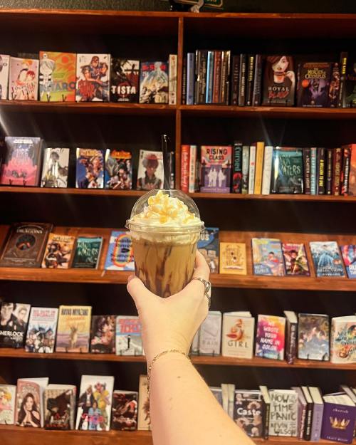 A hand holds a coffee drink topped with whipped cream and caramel, in front of a colorful bookshelf filled with books.