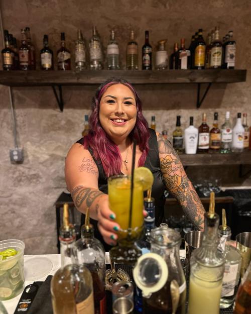 A smiling bartender with colorful hair and tattoos holds a cocktail in front of a well-stocked bar.