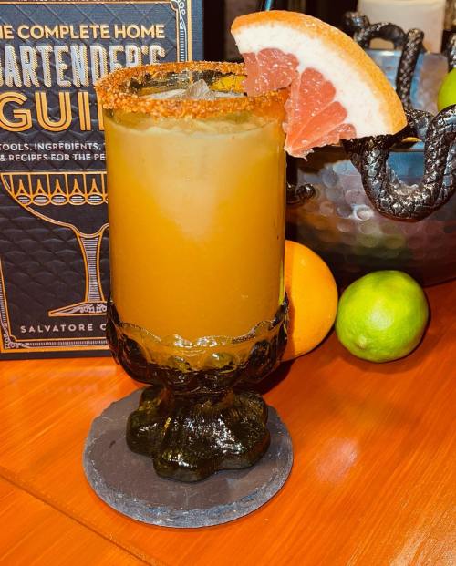 A cocktail in a decorative glass, garnished with a grapefruit slice and rimmed with salt, next to colorful fruits.