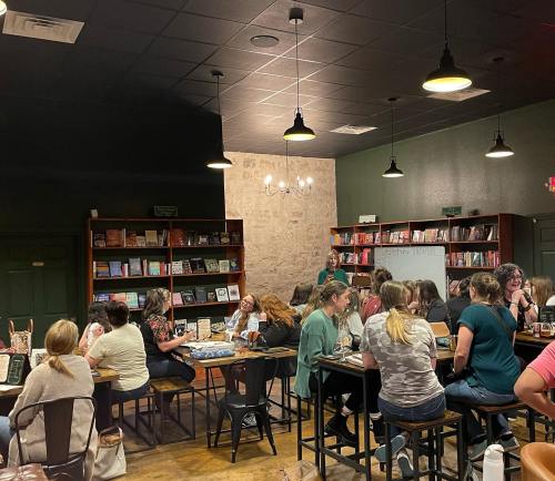 A cozy bookstore café with people seated at tables, engaged in conversation and reading, surrounded by bookshelves.