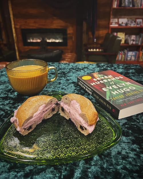 A cozy scene featuring a bagel with cream cheese, a cup of coffee, and a book on a green tablecloth.