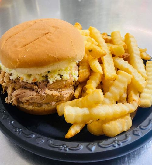 A pulled pork sandwich with coleslaw on a bun, served with a side of crinkle-cut fries on a black plate.