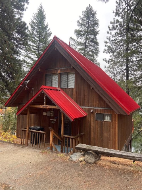 A cozy wooden cabin with a red roof, surrounded by tall trees and a gravel path.