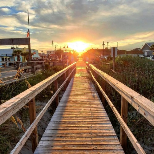 A wooden boardwalk leads towards a sunset, framed by grass and buildings on either side.
