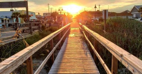 A wooden boardwalk leads towards a sunset, surrounded by grass and buildings, creating a serene coastal scene.