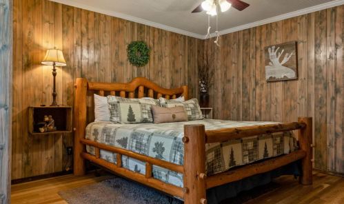 Cozy bedroom with wooden walls, a log bed, decorative pillows, and rustic decor, featuring a lamp and wall art.