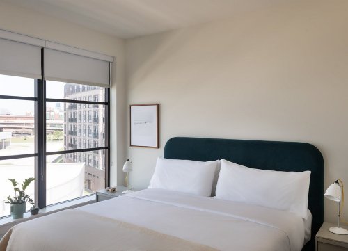 A modern bedroom with a large window, featuring a green upholstered headboard, white bedding, and a small plant on the nightstand.