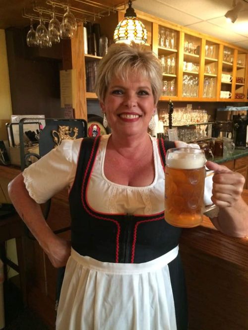 A smiling woman in a traditional dress holds a large beer mug in a cozy bar setting.