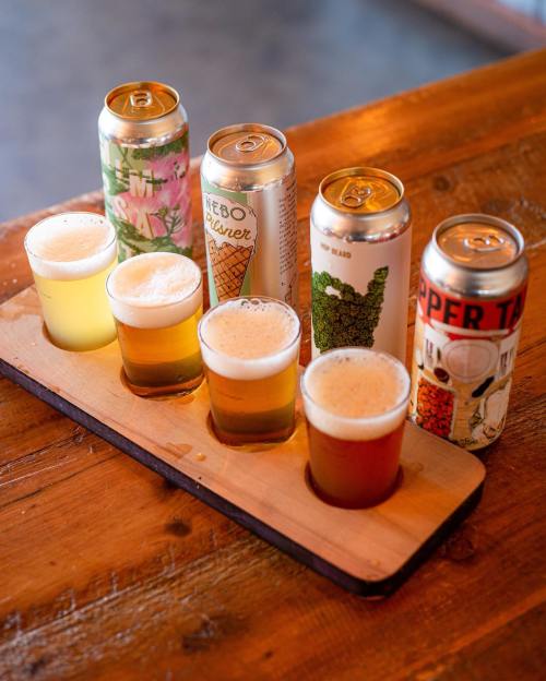 A wooden flight board with three small glasses of beer and four colorful cans on a rustic table.