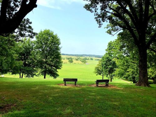 Two benches on a grassy hill overlook a lush green field and trees under a clear blue sky.