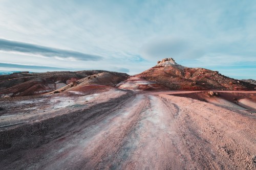 A winding dirt road leads through colorful, rugged terrain under a cloudy sky.