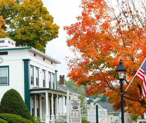 A charming white house with green trim, surrounded by vibrant autumn foliage and an American flag.