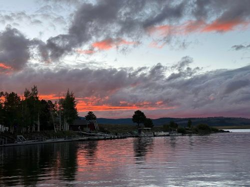 A serene lake at sunset, with colorful clouds reflecting on the water and trees lining the shore.