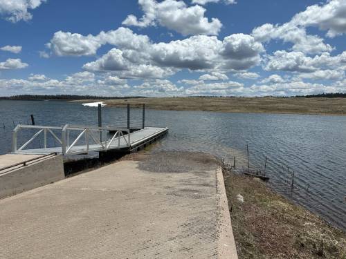 A serene lakeside view featuring a wooden dock, calm water, and fluffy clouds in a blue sky.