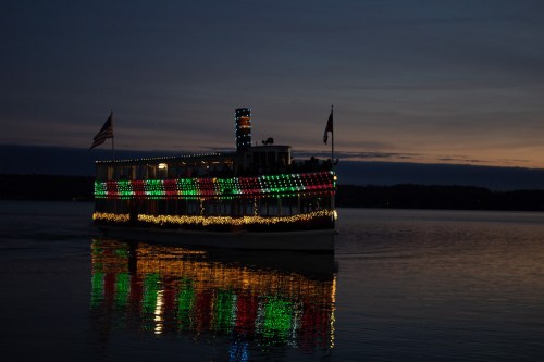 A brightly lit boat with colorful decorations glides across a calm lake at sunset, reflecting on the water.