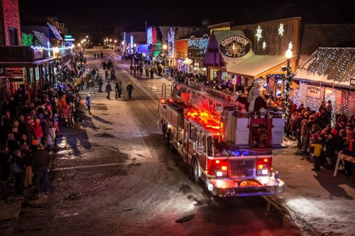 A festive night scene with a fire truck driving through a snowy street lined with holiday lights and a crowd of onlookers.