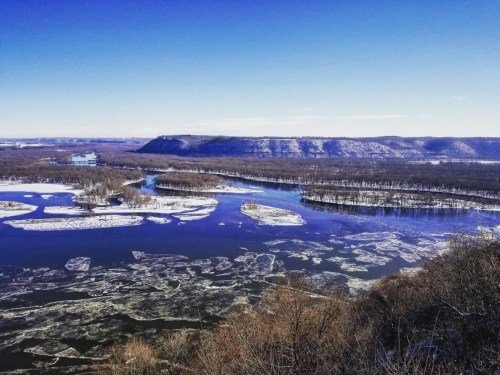 A scenic view of a river with ice floes, surrounded by snow-covered hills under a clear blue sky.