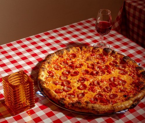 A large pepperoni pizza on a checkered tablecloth, accompanied by a glass of red wine and a stack of orange glasses.