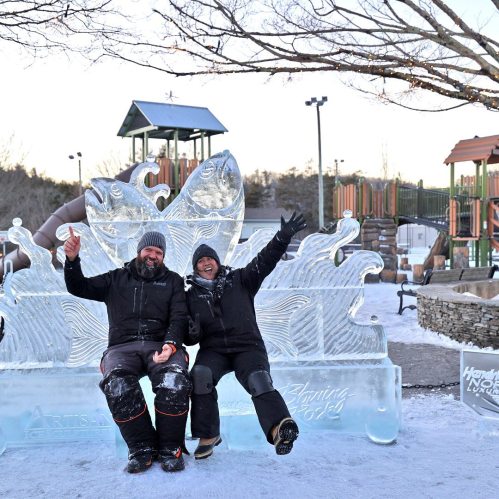 Two people sit on an ice sculpture of fish, smiling and celebrating in a snowy outdoor park.