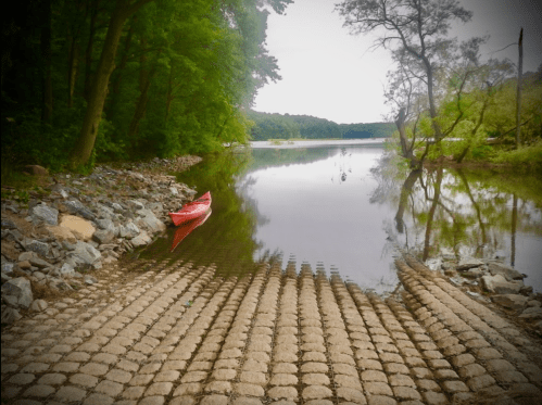 A red kayak rests on a stone ramp by a calm river, surrounded by lush green trees and a serene landscape.