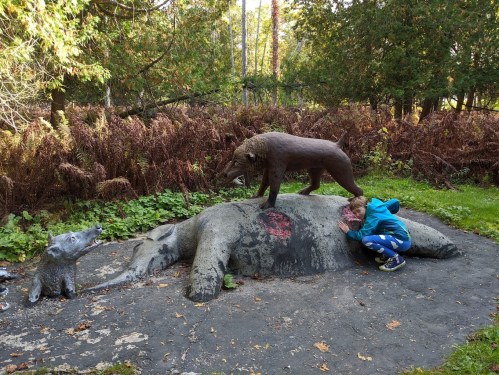 A child interacts with a sculpture of a wild boar standing on a larger animal, surrounded by greenery and autumn foliage.