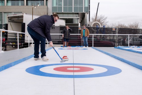 A person curls a stone on an outdoor curling rink while two spectators watch nearby.