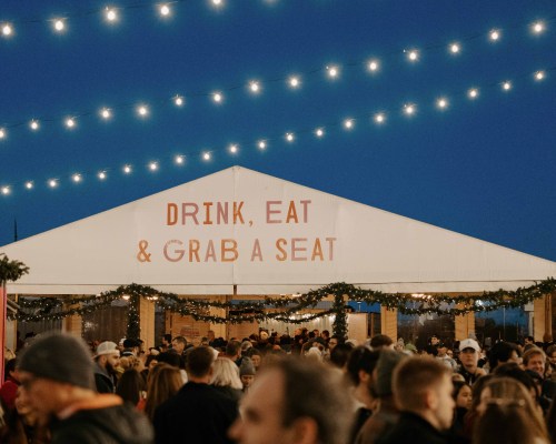 A bustling outdoor event under string lights, featuring a large tent with the words "Drink, Eat & Grab a Seat."