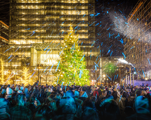 A festive crowd gathers around a brightly lit Christmas tree amidst falling snow in a city square at night.