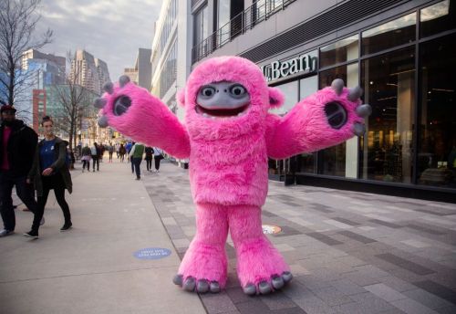 A large, pink furry mascot with big eyes and outstretched arms stands on a city sidewalk, surrounded by people.