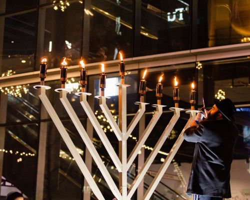 A person lights candles on a large menorah during a festive celebration, with city lights in the background.