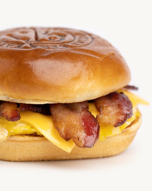 A close-up of a breakfast sandwich with bacon, cheese, and an egg on a soft, toasted bun.