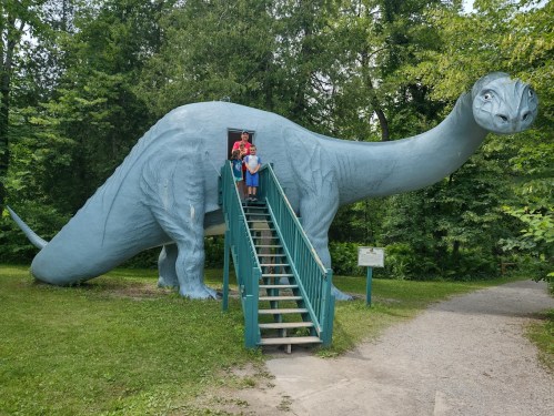 Two children stand on stairs leading into a large blue dinosaur statue in a wooded area.