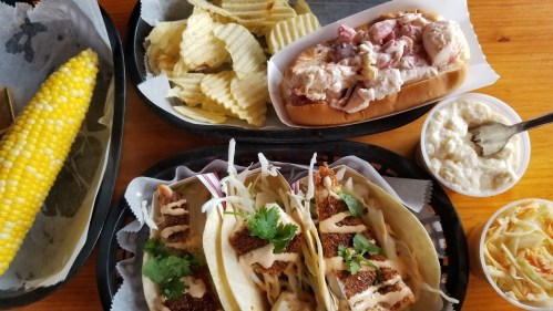 A spread of food featuring tacos, a lobster roll, chips, corn on the cob, and coleslaw in takeout containers.