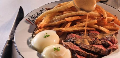 A plate of medium-cooked steak with fries and two poached eggs, topped with a creamy sauce.