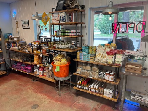 A cozy shop interior featuring wooden shelves filled with various food items, snacks, and local products. A neon "OPEN" sign is visible.