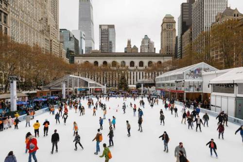 A bustling ice skating rink in a city park, surrounded by tall buildings and trees, with many skaters enjoying the activity.