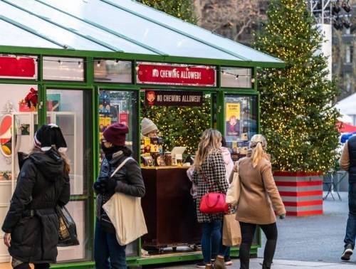 A festive market stall with holiday decorations and shoppers in winter attire, surrounded by Christmas trees.