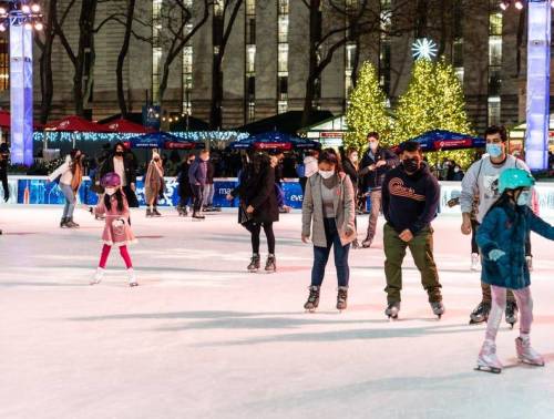 People ice skating at a festive outdoor rink, with holiday lights and a decorated tree in the background.