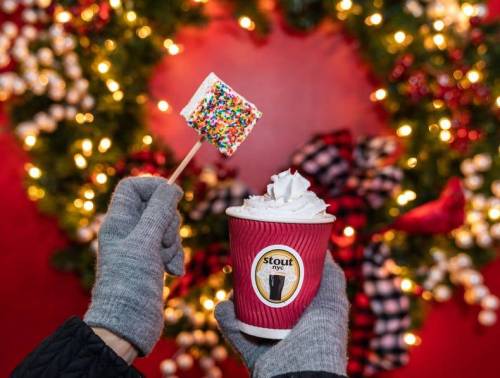 A hand in a gray glove holds a festive cup of hot chocolate topped with whipped cream and a colorful marshmallow on a stick.