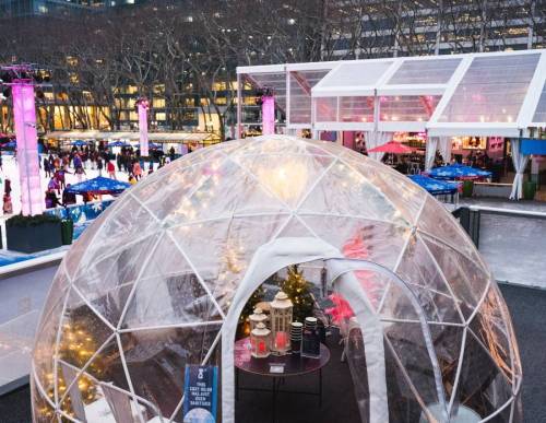 A transparent igloo structure with lights, surrounded by a festive outdoor market and ice skating rink.