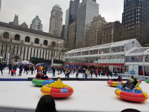 People ice skating in colorful bumper cars at a winter event in a city park, surrounded by tall buildings.