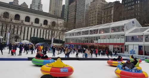 Colorful bumper cars on ice with people skating and a large building in the background, set in a cityscape.