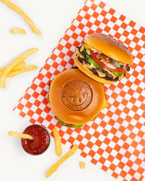 Two delicious burgers on a checkered paper with fries and a small cup of ketchup.
