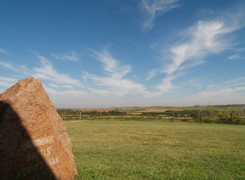 A stone marker in a grassy field, with a blue sky and wispy clouds above, overlooking distant hills and a wooden fence.