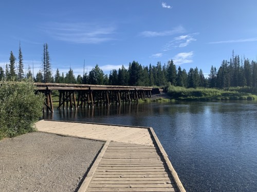 A wooden dock extends into a calm river, surrounded by lush greenery and tall trees under a clear blue sky.